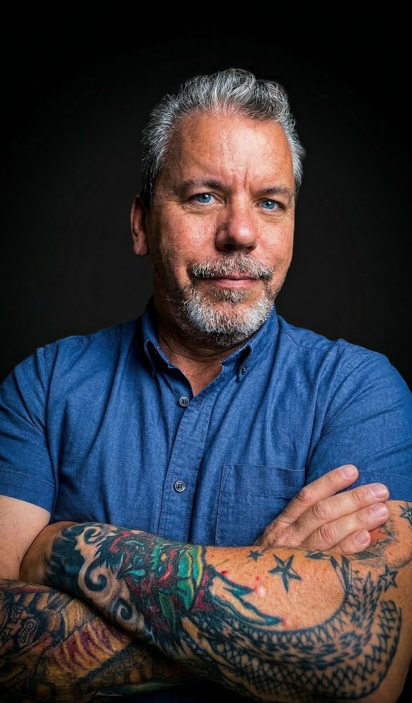 Michael Sanford, arms crossed, in a blue shirt with tattooed forearms, against a dark background